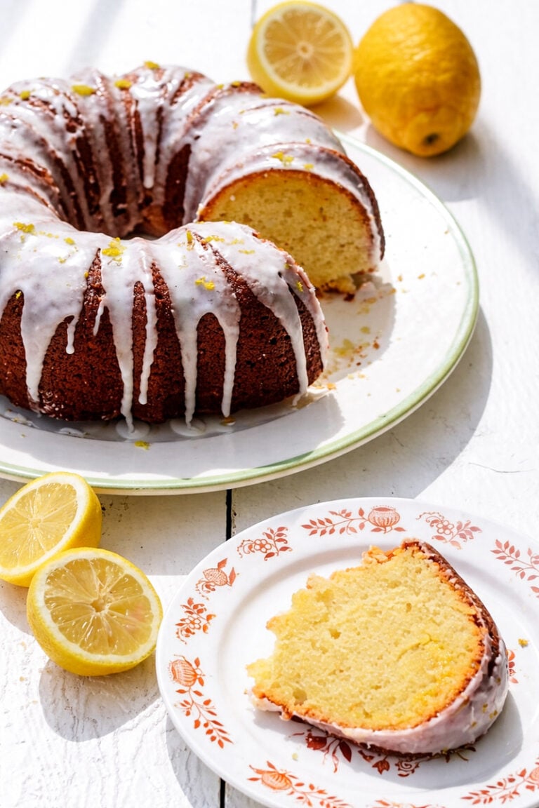 A lemon curd cake bundt with white glaze sits on a plate, with a slice served on a floral-patterned dish. Fresh lemons, some halved, are scattered nearby on a white surface.