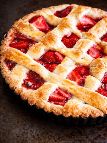 A close-up of a freshly baked strawberry crostata with a golden brown lattice crust, showing juicy red strawberries beneath the woven pastry. The pie sits in a fluted tart pan on a dark surface.