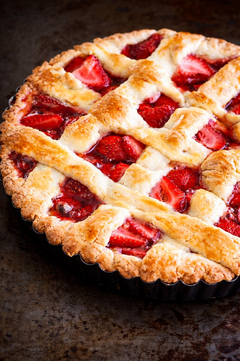 A close-up of a freshly baked strawberry crostata with a golden brown lattice crust, showing juicy red strawberries beneath the woven pastry. The pie sits in a fluted tart pan on a dark surface.