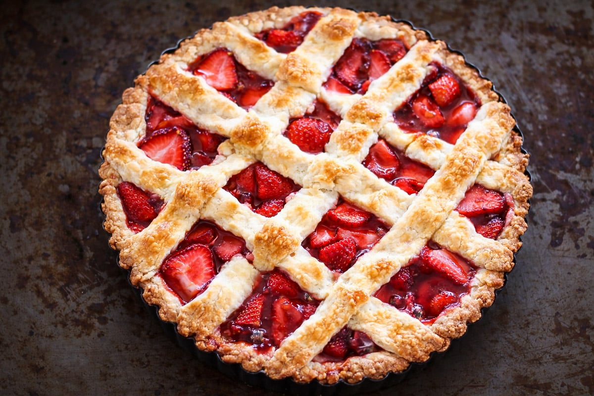 A freshly baked Strawberry Crostata pie with a golden, flaky lattice crust, showing juicy red strawberries in the filling, sits on a dark rustic surface.