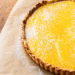 A lemon curd tart with a golden yellow filling and lightly dusted powdered sugar sits on brown parchment paper on a wooden surface.