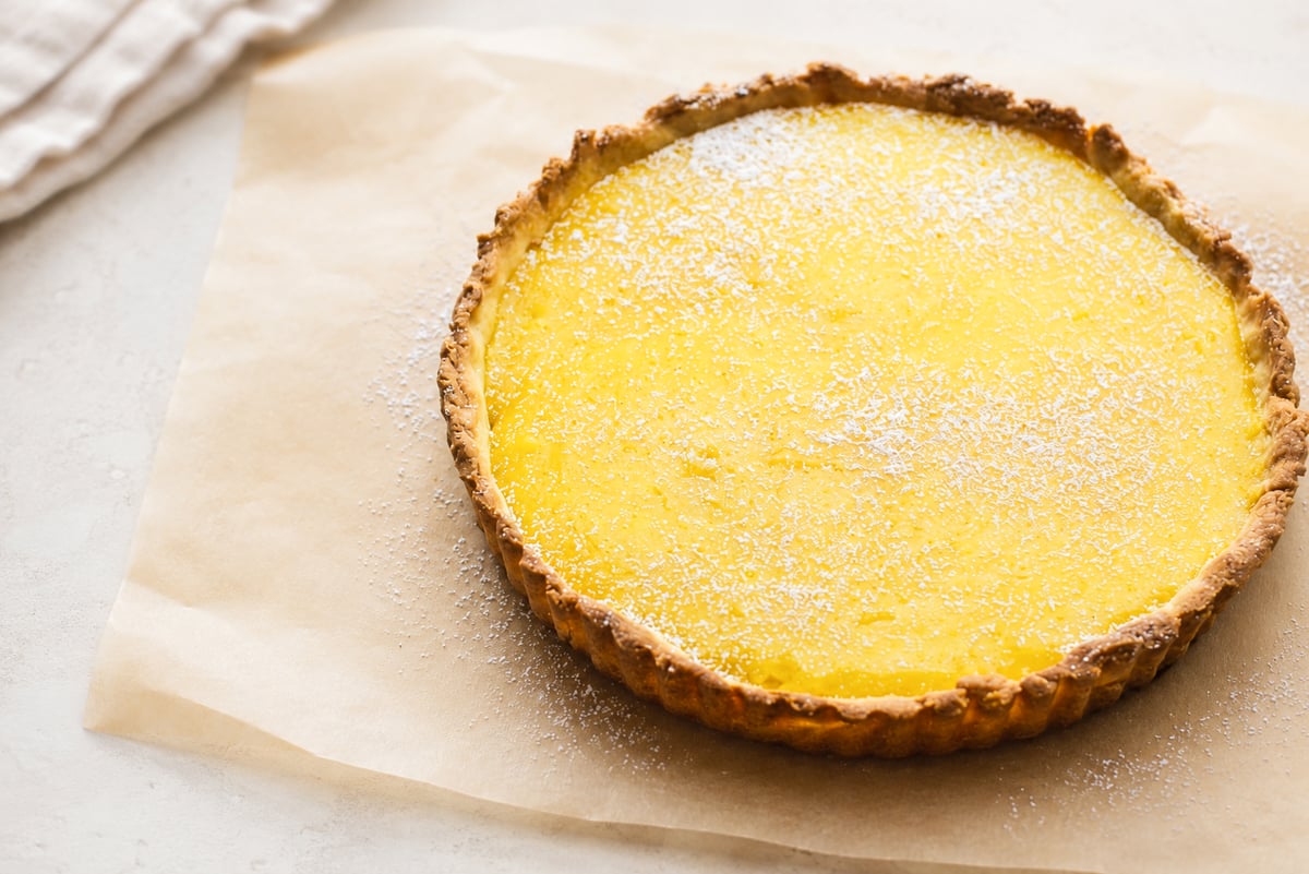 A freshly baked lemon curd tart with a golden crust sits on parchment paper, lightly dusted with powdered sugar.