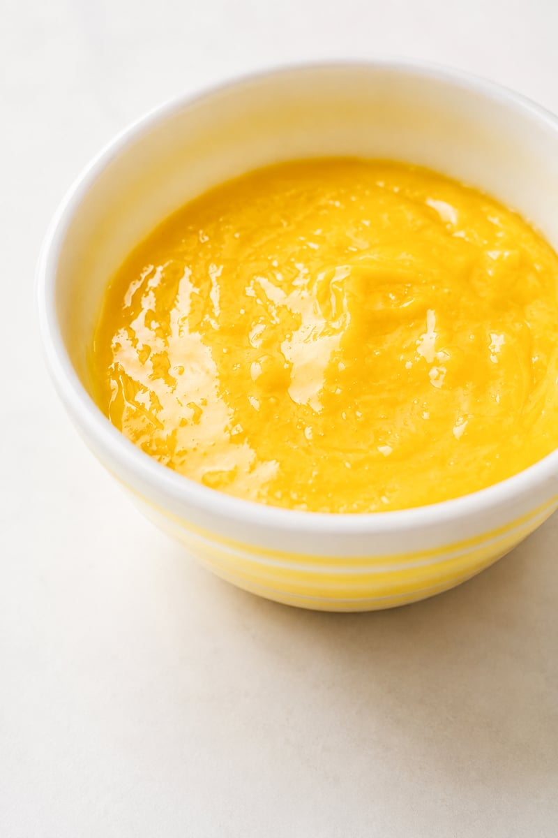 A white bowl filled with smooth, bright yellow lemon curd, reminiscent of the vibrant filling in a classic lemon curd tart, is placed on a light-colored surface.