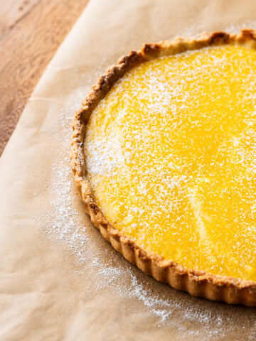 A lemon curd tart with a golden yellow filling and lightly dusted powdered sugar sits on brown parchment paper on a wooden surface.