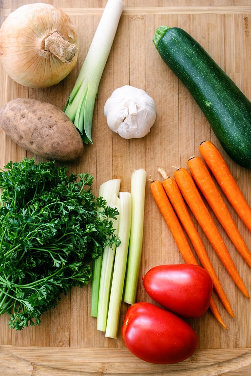 Spring Minestrone A variety of fresh vegetables for Spring Minestrone sit on a wooden cutting board, including an onion, leek, zucchini, garlic, potato, bunch of parsley, celery stalks, carrots, and two Roma tomatoes.