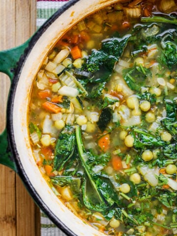 A close-up of a pot filled with Spring Minestrone, featuring leafy greens, chickpeas, diced carrots, celery, onions, and herbs, sitting on a wooden surface with a striped cloth underneath.