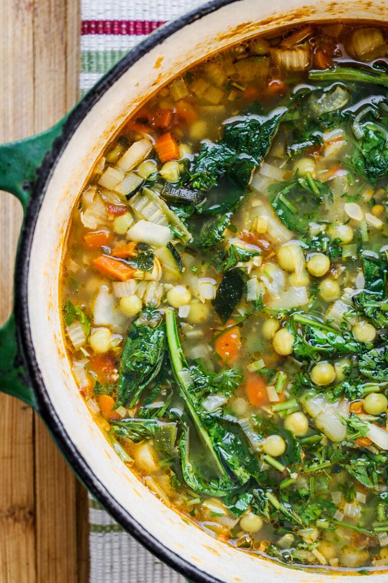 A close-up of a pot filled with Spring Minestrone, featuring leafy greens, chickpeas, diced carrots, celery, onions, and herbs, sitting on a wooden surface with a striped cloth underneath.