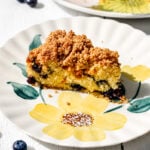 A slice of Blueberry Mango Buckle sits on a floral-patterned plate, with fresh blueberries scattered on a white surface and part of the remaining cake visible in the background.