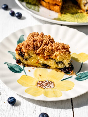 A slice of Blueberry Mango Buckle sits on a floral-patterned plate, with fresh blueberries scattered on a white surface and part of the remaining cake visible in the background.