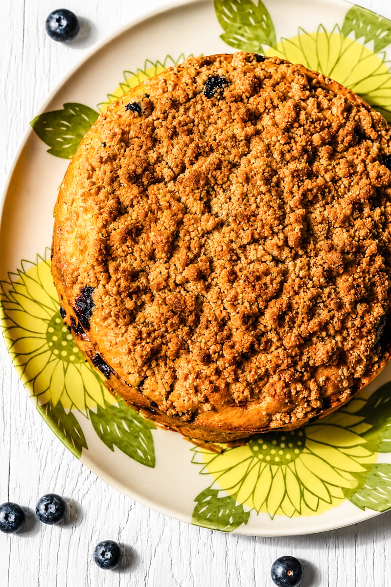 A round Blueberry Mango Buckle crumb cake sits on a plate with yellow sunflower designs, with a few fresh blueberries scattered on a white wooden surface nearby.