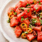 A plate of Asian Watermelon Salad featuring watermelon cubes topped with sliced jalapeños, chopped cilantro, and crispy shallots, all beautifully arranged on a white ceramic dish.