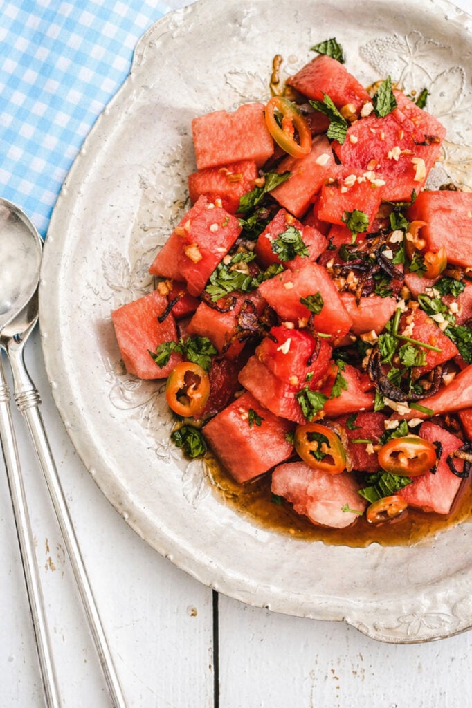 A plate of Asian Watermelon Salad with fresh cilantro, sliced chili peppers, and chopped onions in a light dressing, served on a rustic white dish beside two silver spoons on a blue and white checkered napkin.