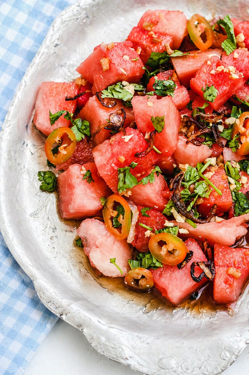 A silver plate filled with Asian Watermelon Salad—cubed watermelon, garnished with sliced jalapeños, chopped cilantro, and caramelized onions—sits on a blue and white checkered cloth.