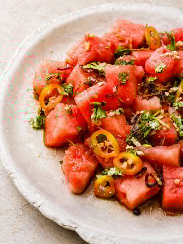 A plate of Asian Watermelon Salad featuring watermelon cubes topped with sliced jalapeños, chopped cilantro, and crispy shallots, all beautifully arranged on a white ceramic dish.