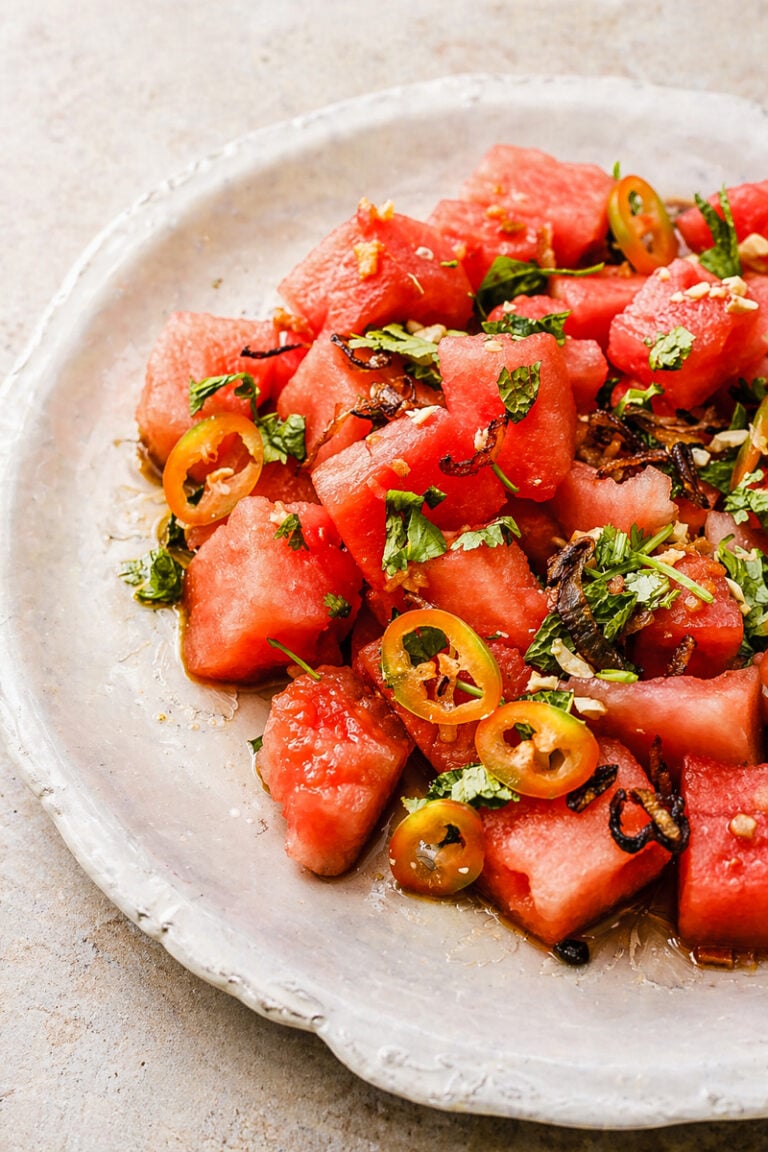 A plate of Asian Watermelon Salad featuring watermelon cubes topped with sliced jalapeños, chopped cilantro, and crispy shallots, all beautifully arranged on a white ceramic dish.