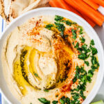 A bowl of Easy Homemade Hummus topped with olive oil, paprika, and chopped parsley, served with pita bread, carrot sticks, and cucumber slices.