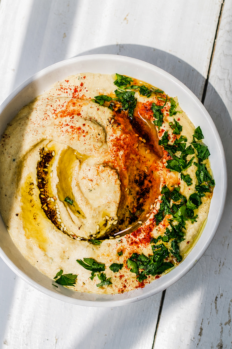 A bowl of easy homemade hummus topped with olive oil, chopped parsley, and paprika sits on a white wooden surface, with sunlight casting soft shadows.