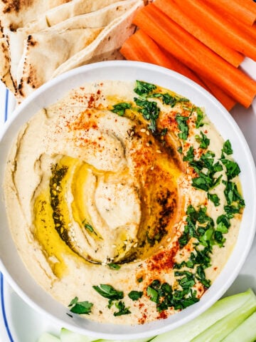 A bowl of Easy Homemade Hummus topped with olive oil, paprika, and chopped parsley, served with pita bread, carrot sticks, and cucumber slices.