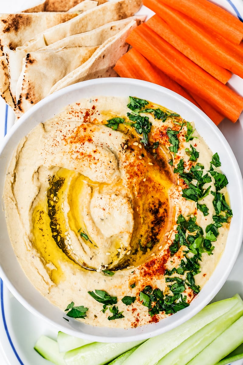 A bowl of Easy Homemade Hummus topped with olive oil, paprika, and chopped parsley, served with pita bread, carrot sticks, and cucumber slices.