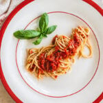 A plate of Spaghetti with Fresh Tomato Sauce, garnished with basil leaves, sits on a white plate with a red rim. A glass of water and a beige napkin are nearby on a light colored surface.