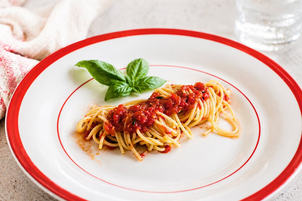 A small serving of Spaghetti with Fresh Tomato Sauce, garnished with basil, sits on a white plate with a red rim. A glass of water and a cloth napkin are in the background.