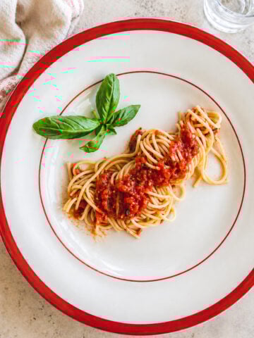 A plate of Spaghetti with Fresh Tomato Sauce, garnished with basil leaves, sits on a white plate with a red rim. A glass of water and a beige napkin are nearby on a light colored surface.