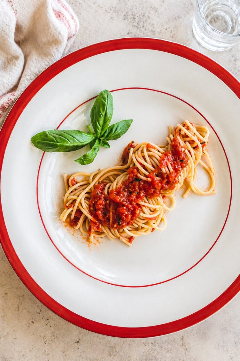 A plate of Spaghetti with Fresh Tomato Sauce, garnished with basil leaves, sits on a white plate with a red rim. A glass of water and a beige napkin are nearby on a light colored surface.