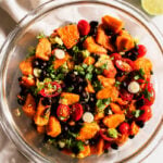A glass bowl filled with a vibrant papaya black bean salad featuring sweet potatoes, cherry tomatoes, chopped green onions, and cilantro, with two lime halves and a white cloth nearby on a light surface.