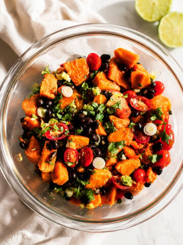 A glass bowl filled with a vibrant papaya black bean salad featuring sweet potatoes, cherry tomatoes, chopped green onions, and cilantro, with two lime halves and a white cloth nearby on a light surface.