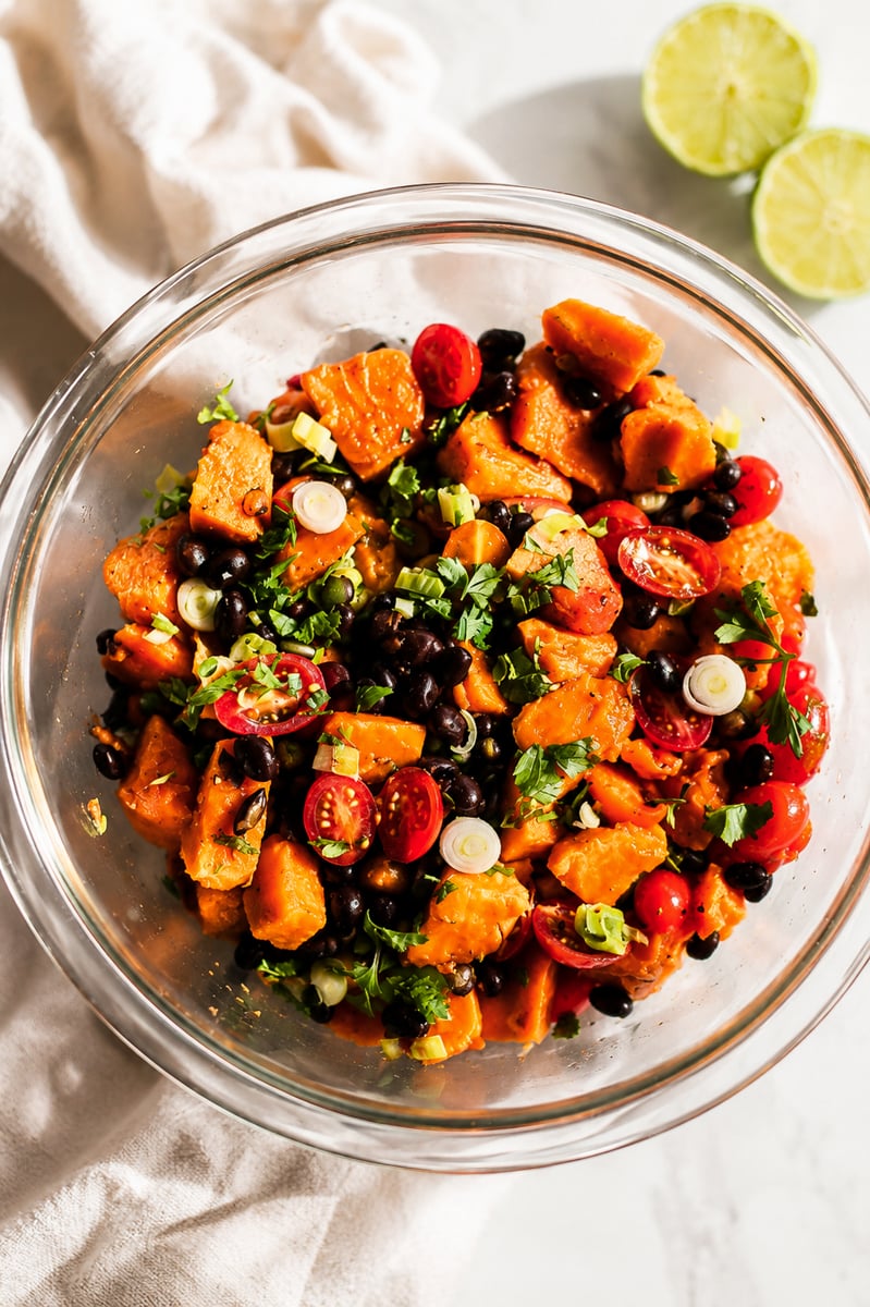 A glass bowl filled with a vibrant papaya black bean salad featuring sweet potatoes, cherry tomatoes, chopped green onions, and cilantro, with two lime halves and a white cloth nearby on a light surface.