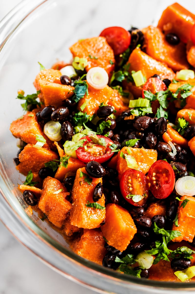 A glass bowl filled with a vibrant papaya black bean salad featuring roasted sweet potato cubes, halved cherry tomatoes, chopped green onions, and fresh cilantro.