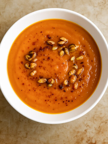 A white bowl of creamy Roasted Butternut Squash Soup, garnished with roasted seeds and a sprinkle of red spices, sits on a light brown surface.