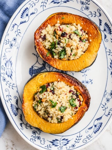 Two halves of a stuffed acorn squash with quinoa, cranberries, pine nuts, and herbs, served on a blue and white patterned platter.
