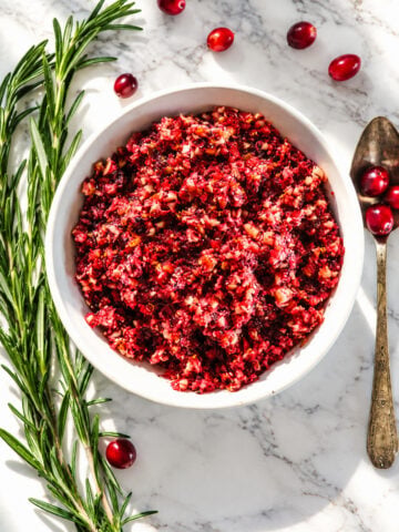 A white bowl filled with cranberry relish, accented by fresh rosemary sprigs and a vintage spoon on a marble surface.