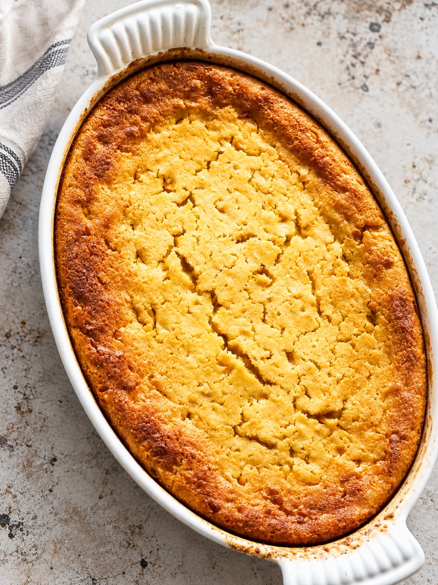 Golden brown Sweet Corn Spoonbread baked in an oval white ceramic dish, resting on a light, textured surface next to a striped cloth. The top is cracked and crisp.