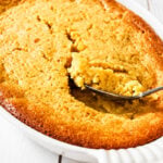 A golden brown Sweet Corn Spoonbread casserole in a white baking dish, with a spoon scooping out a portion. The cornbread looks moist and fluffy inside, with a lightly crisped edge.
