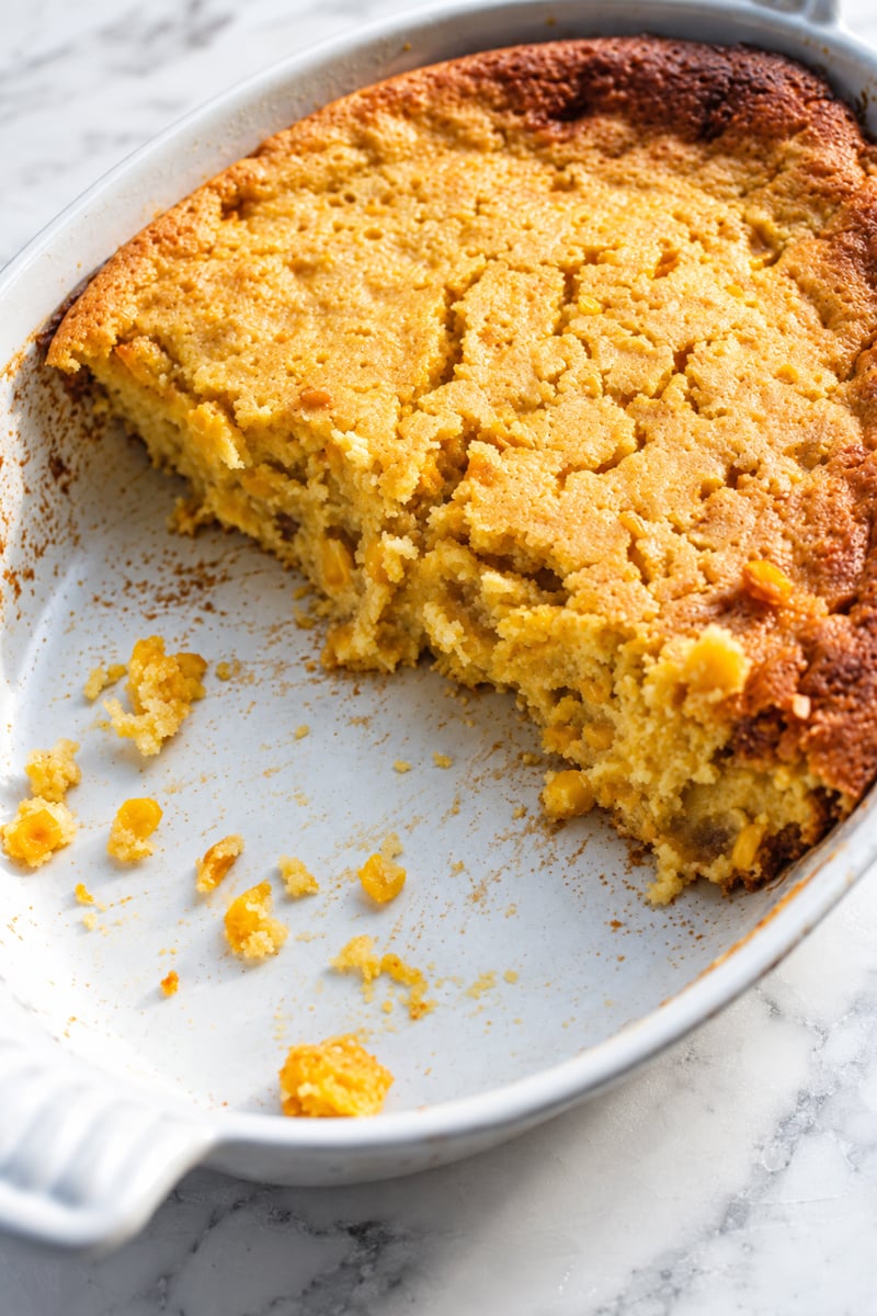 A partially eaten Sweet Corn Spoonbread casserole in a white oval baking dish on a marble surface. The golden brown top is slightly cracked, and crumbs are scattered in the dish.