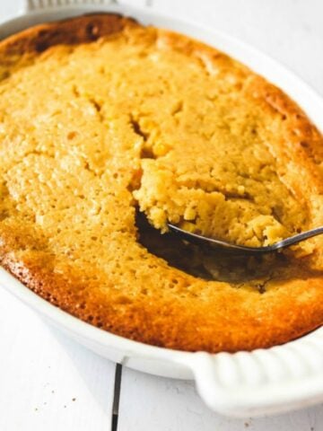 A spoon scooping into a golden-brown Sweet Corn Spoonbread casserole, showing a moist, crumbly interior in a white oval dish on a white surface.