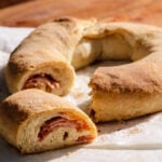A golden-brown Casatiello, the classic ring-shaped bread, is sliced to reveal layers of ham and cheese inside, resting on parchment paper atop a wooden surface.