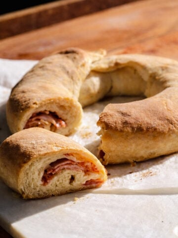 A golden-brown Casatiello, the classic ring-shaped bread, is sliced to reveal layers of ham and cheese inside, resting on parchment paper atop a wooden surface.