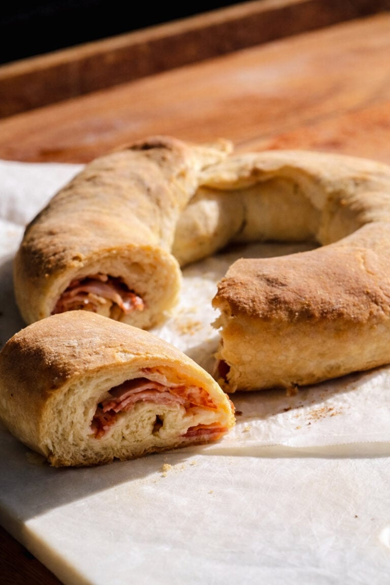 A golden-brown Casatiello, the classic ring-shaped bread, is sliced to reveal layers of ham and cheese inside, resting on parchment paper atop a wooden surface.