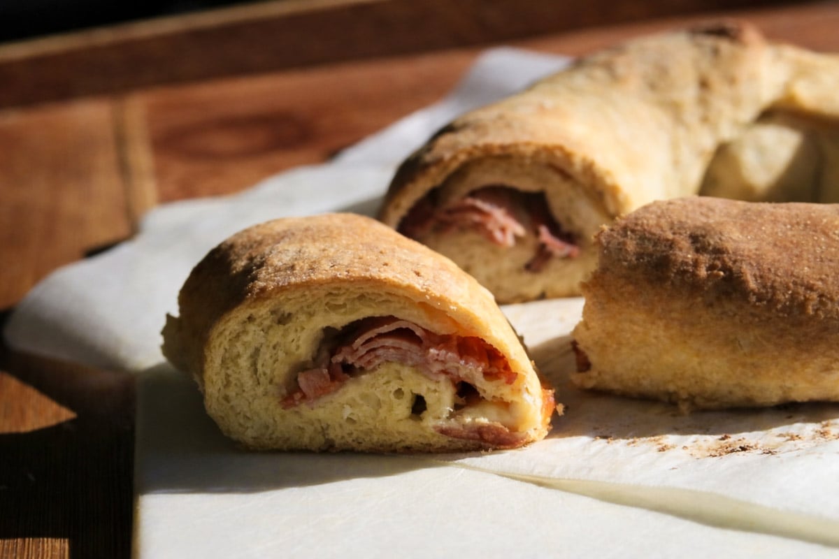 A close-up of a sliced, golden-brown Casatiello bread roll filled with layers of sliced meat, resting on parchment paper on a wooden surface. Sunlight highlights the rich texture of the bread and savory filling.