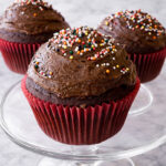Three One-Bowl Chocolate Cupcakes with chocolate frosting and colorful sprinkles sit on a glass cake stand. The cupcakes are in red paper liners and displayed on a light gray marble surface.