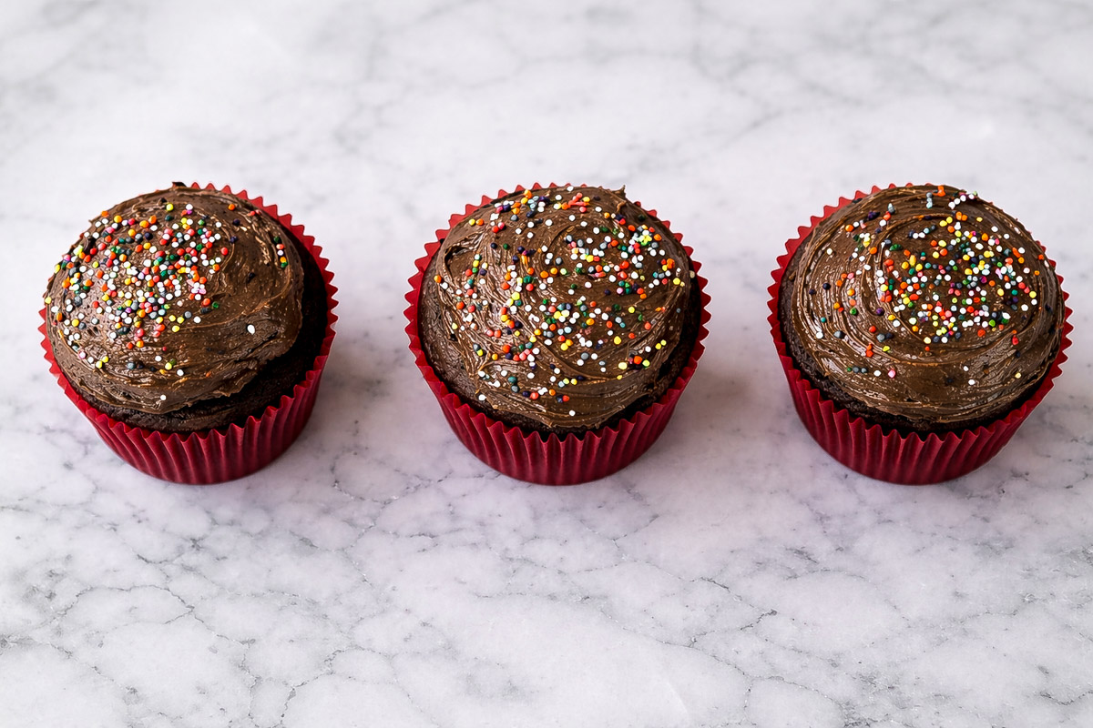 Three One-Bowl Chocolate Cupcakes with Chocolate Frosting in red paper liners are arranged in a row on a white marble surface, each topped with colorful round sprinkles.