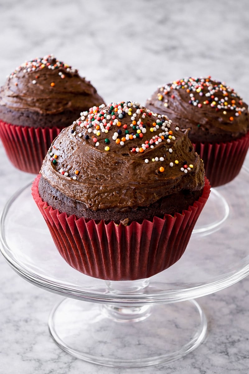 Three One-Bowl Chocolate Cupcakes with chocolate frosting and colorful sprinkles sit on a glass cake stand. The cupcakes are in red paper liners and displayed on a light gray marble surface.