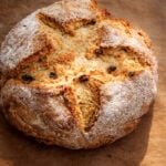 A round loaf of Irish Soda Bread-Spotted Dog with a golden crust, lightly dusted with flour, featuring a deep cross cut on top and visible raisins, rests on brown parchment paper.