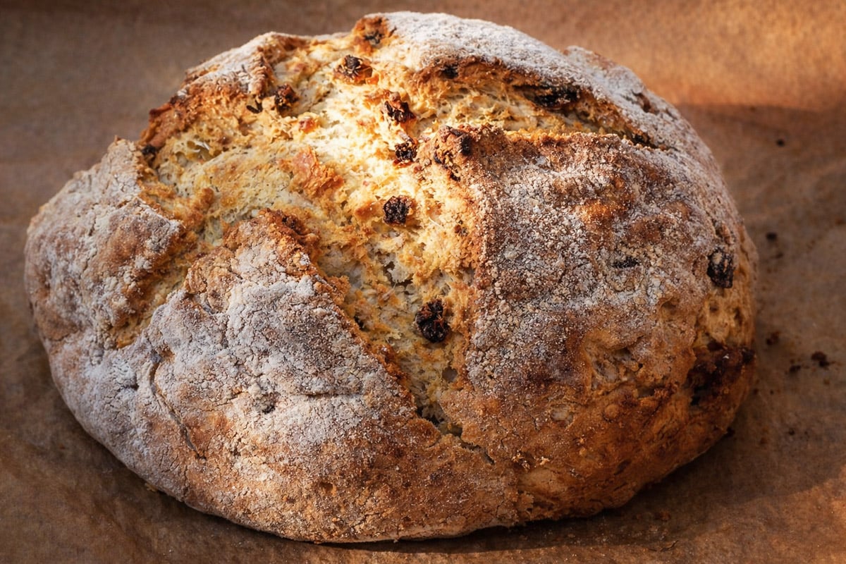 A round loaf of Irish Soda Bread-Spotted Dog with a golden-brown crust, dusted with flour and dotted with raisins, sits on a sheet of brown parchment paper. Sunlight highlights its cracked surface.