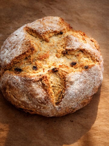 A round loaf of Irish Soda Bread-Spotted Dog with a golden crust, lightly dusted with flour, featuring a deep cross cut on top and visible raisins, rests on brown parchment paper.