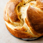 A round loaf of golden-brown, fluffy Cinnamon Raisin Round Challah Bread with a cracked top sits on a light surface, showcasing its soft, airy texture and shiny crust.