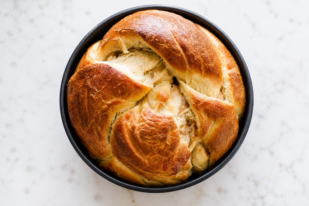 A Cinnamon Raisin Round Challah Bread with a golden-brown crust sits in a black baking pan on a white, speckled surface. The loaf’s signature braided design hints at its tender, flavorful interior swirled with sweet cinnamon and plump raisins.